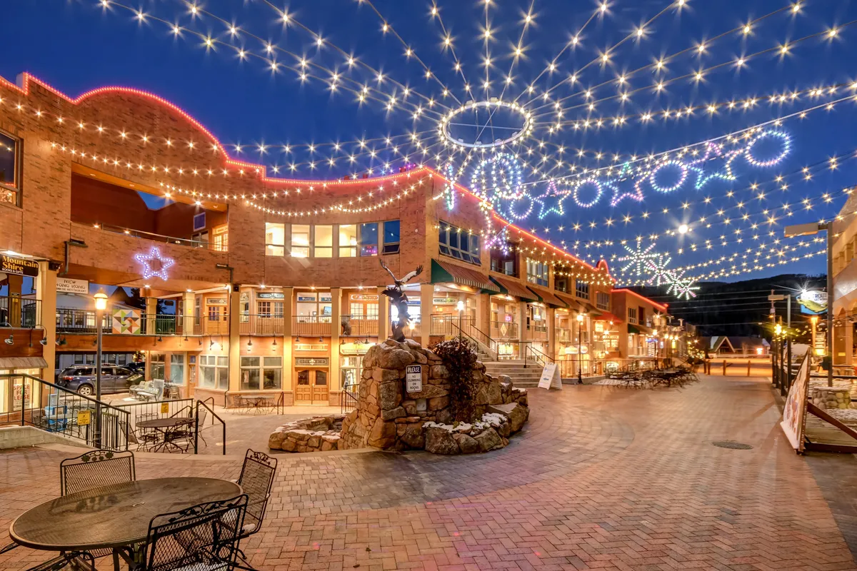 An inviting exterior scene featuring twinkling fairy lights, decorative stars, a central fountain, and outdoor seating areas surrounded by a vibrant atmosphere. The image conveys a lively local scene of dining, shopping, and festive events.