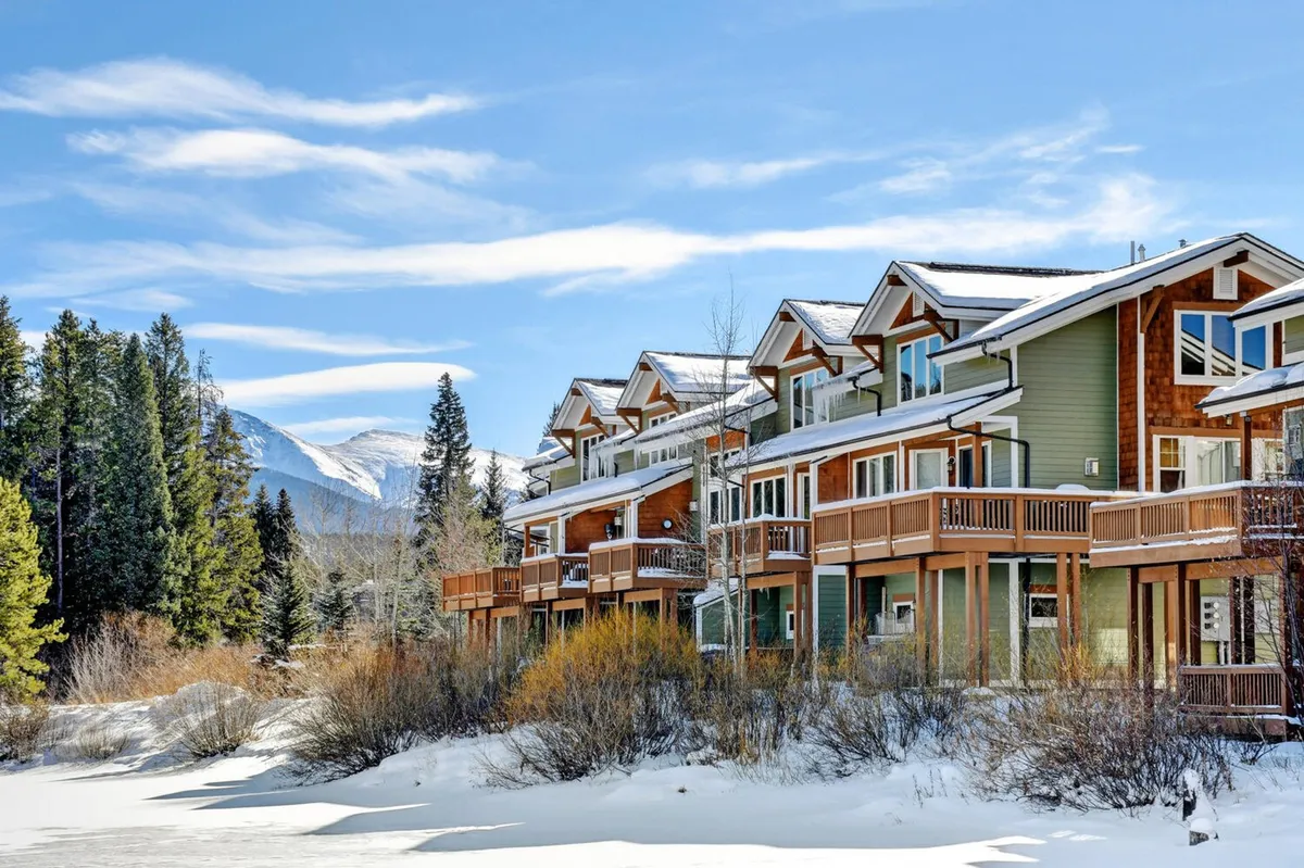 The image showcases a stunning snow-covered luxury villa set against the breathtaking backdrop of towering Colorado mountains. Lush evergreen trees frame the property while a bright blue sky bathes the scene in tranquil light. The villa's balconies invite relaxation, and surrounding wilderness hints at endless outdoor adventures like skiing, snowboarding, and hiking.