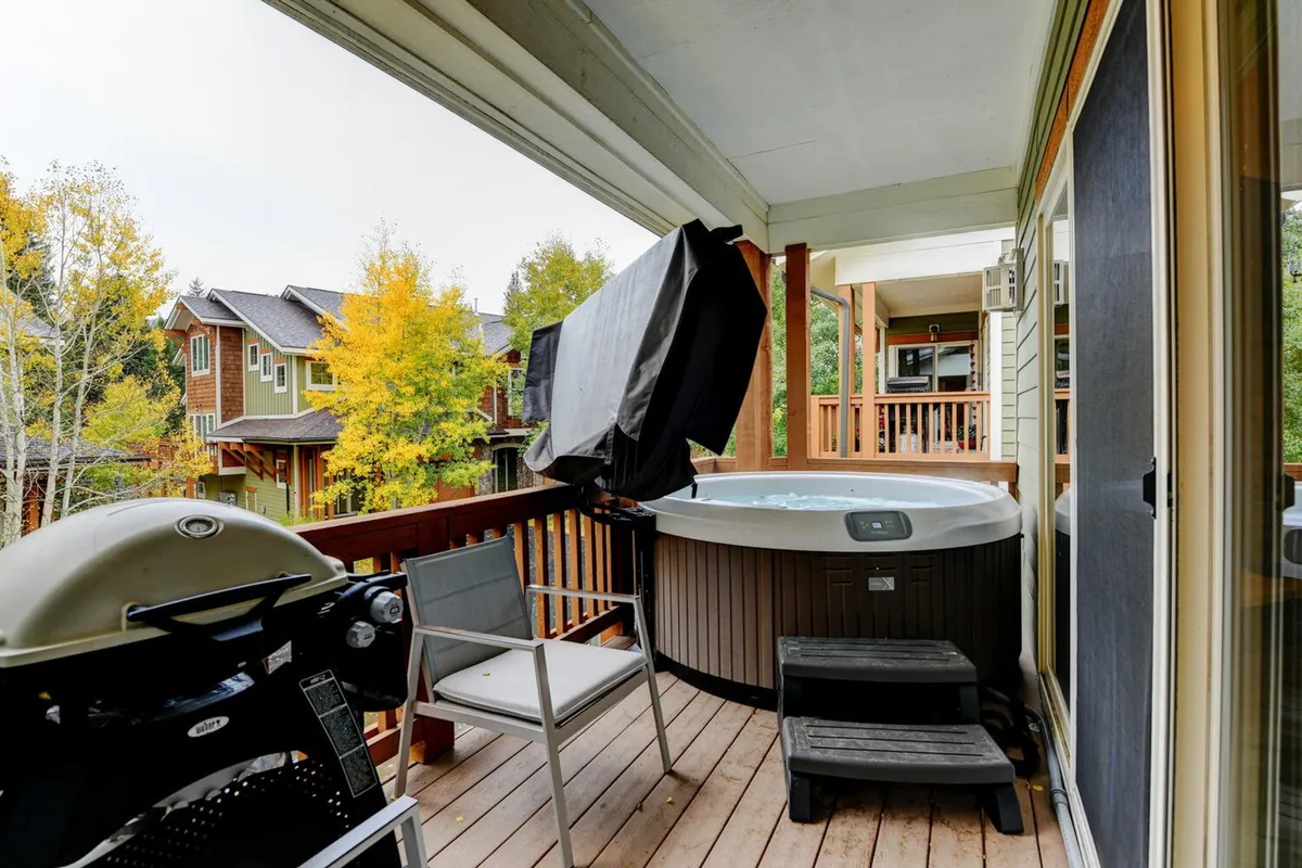 This image features a private wooden deck surrounded by lush trees, with a bubbling hot tub and a gas grill prominently displayed. The setup also includes ample space for outdoor lounging, conveying a sense of tranquility and immersion in nature.