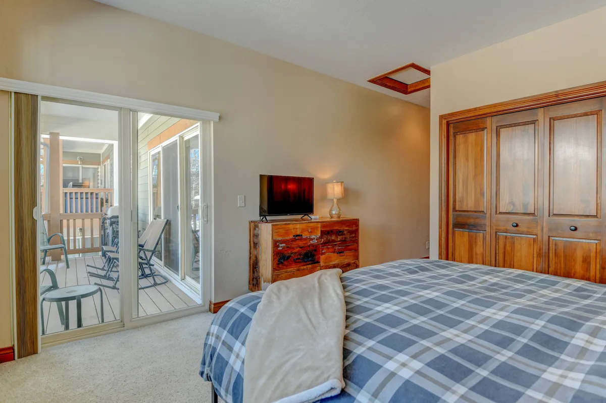 A cozy bedroom featuring a bed with plaid bedding, a wooden dresser, and sliding glass doors that open onto the balcony. The doors provide a direct visual link between the comfortable indoor setting and the inviting outdoor space beyond.